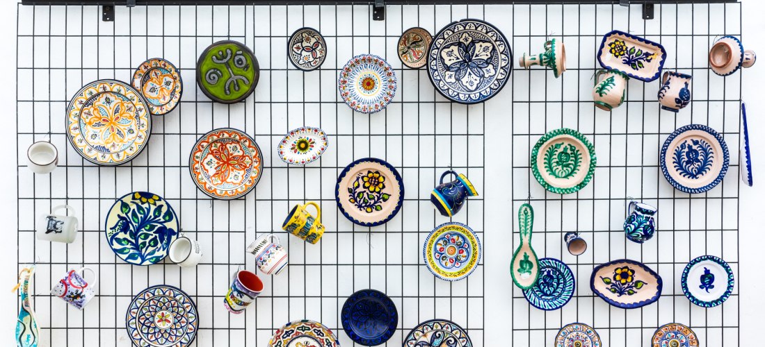 Bunte, handbemalte Keramikteller, Tassen und Schalen hängen dekorativ an einem Gitter vor weißer Wand. Die Muster sind vielfältig, meist floral oder geometrisch