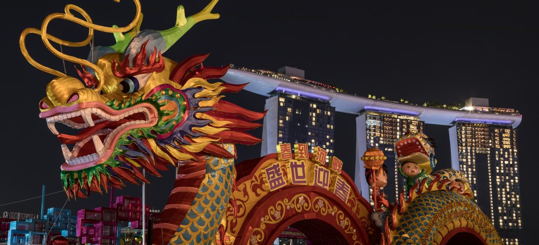 Farbenfrohe Drachenfigur mit großen Zähnen und bunten Schuppen im Vordergrund. Dahinter moderne Hochhäuser bei Nacht, darunter das beleuchtete Marina Bay Sands Hotel in Singapur. Die Szene zeigt eine festliche Dekoration zum chinesischen Neujahrsfest.