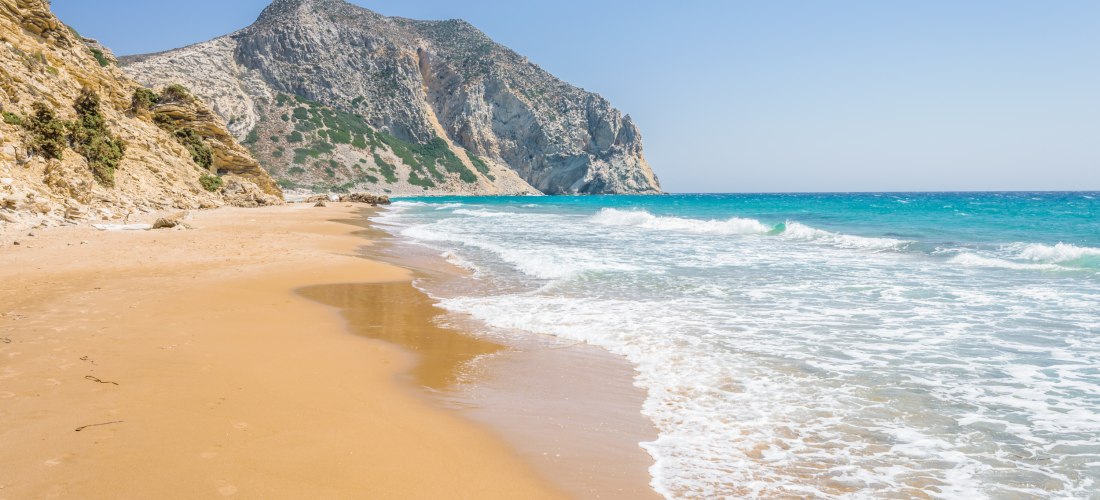 Weitläufiger, menschenleerer Sandstrand mit goldfarbenem Sand und sanften Wellen, die an das Ufer rollen. Im Hintergrund ragt ein steiler, felsiger Berg empor. Die Landschaft wirkt ursprünglich und naturbelassen, das Wasser schimmert türkis und der Himmel