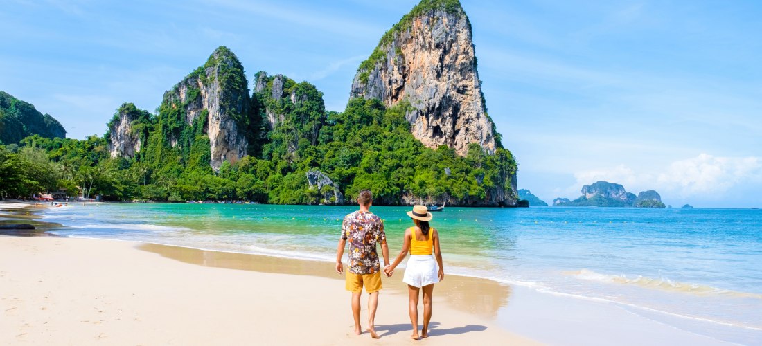 Railay Beach in Krabi, Thailand: Paar spaziert am weißen Sandstrand vor türkisfarbener Andamanensee und imposanten Kalksteinfelsen.
