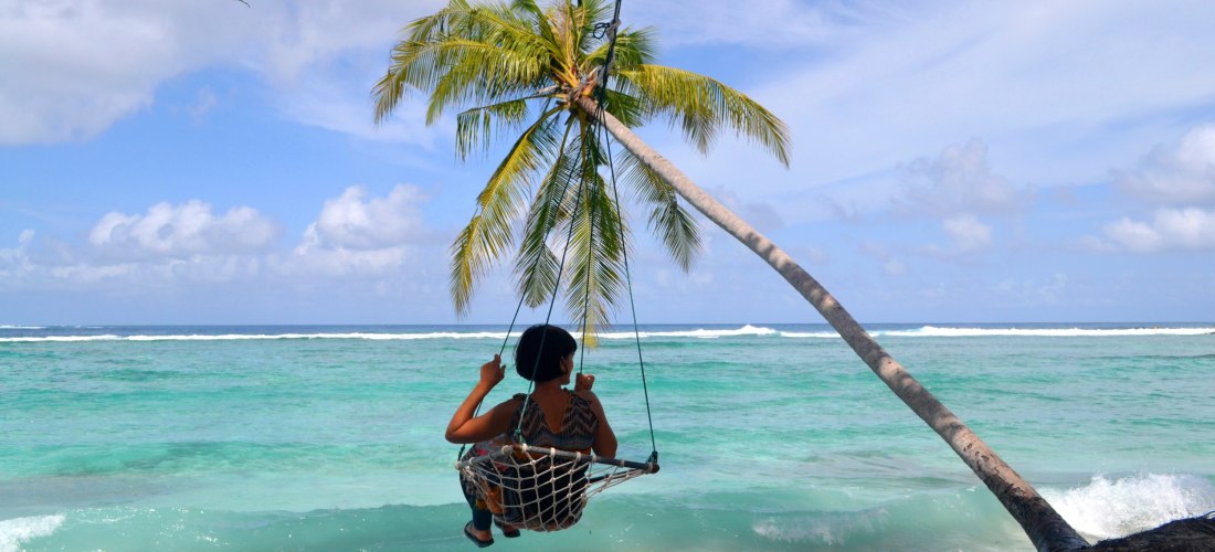 Person sitzt auf einer Schaukel unter einer Palme direkt am Strand der Malediven mit Blick auf das türkisblaue Meer