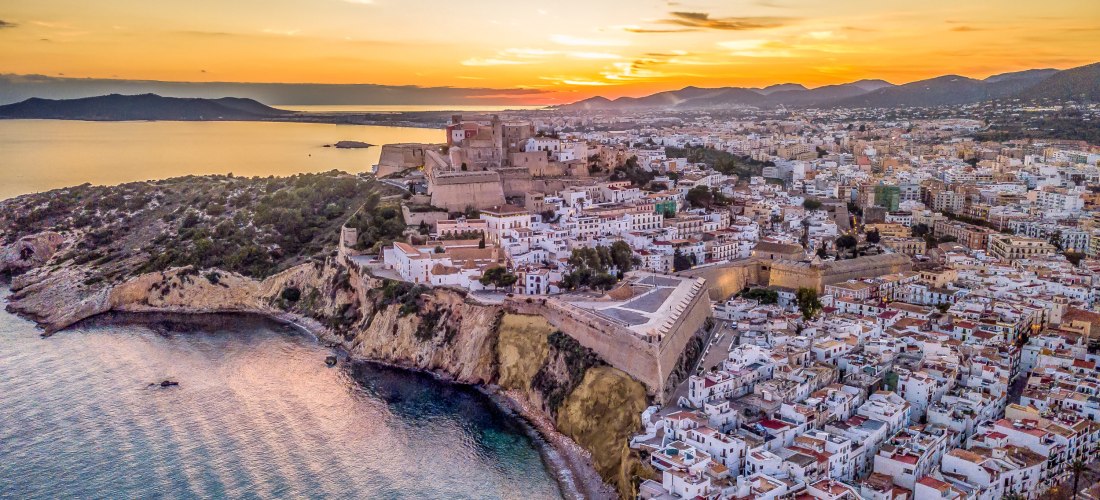 Luftaufnahme von Ibiza-Stadt mit der historischen Festung Dalt Vila auf einem Hügel über dem Meer. Weiße Häuser ziehen sich entlang der Küstenlinie, im Hintergrund breiten sich Stadt und Berge unter einem farbenfrohen Sonnenuntergang aus