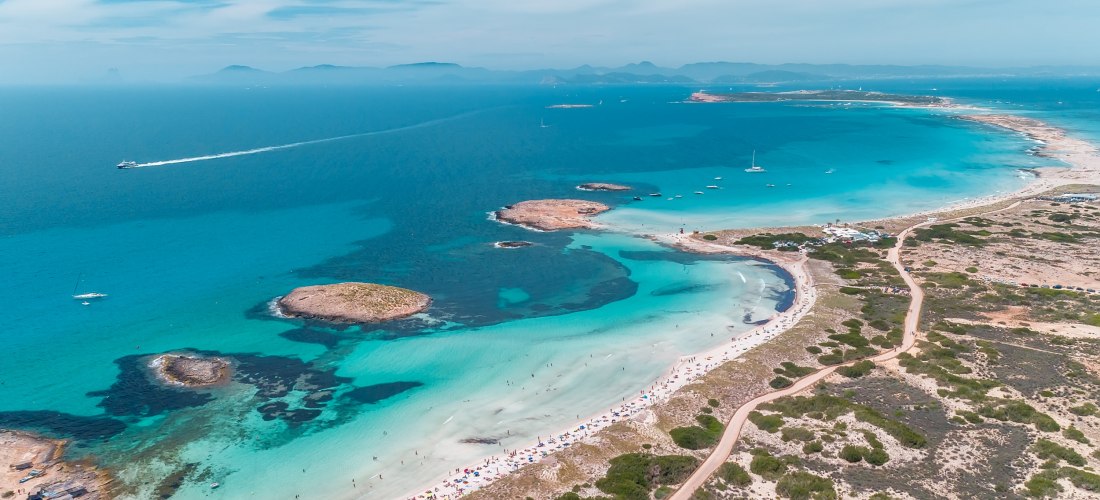 Luftaufnahme eines weit geschwungenen, weißen Sandstrandes auf Formentera mit türkisblauem Wasser und kleinen vorgelagerten Felsen. Zahlreiche Sonnenschirme säumen den Strand, im Hintergrund ziehen Boote durch das klare Meer. Die Küste ist von Dünen und n