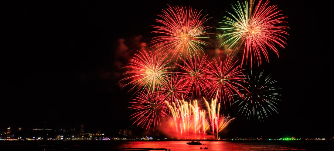 Farbenfrohes Feuerwerk in Rot und Grün über dem Meer in Pattaya. Menschen stehen am Strand und beobachten das Lichtspektakel.