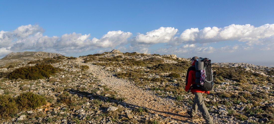 Das Bild zeigt einen Wanderweg in einer felsigen Berglandschaft bei strahlend blauem Himmel. Im Vordergrund ist ein einzelner Wanderer mit roter Jacke, grauer Hose und einem großen schwarzen Rucksack zu sehen. Er geht auf einem schmalen, steinigen Pfad, d