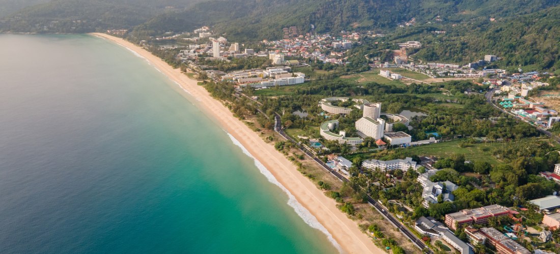 Luftaufnahme vom Karon Beach auf Phuket mit goldenem Sandstrand, türkisfarbenem Wasser und grünen Hügeln im Hintergrund – einer der schönsten Strände Thailands.
