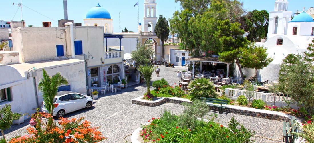 Megalochori auf Santorini: blumengeschmückter Dorfplatz mit weiß gekalkten Häusern, blauem Kuppeldach und Kirchturm-Bogen; Tavernen unter Bäumen