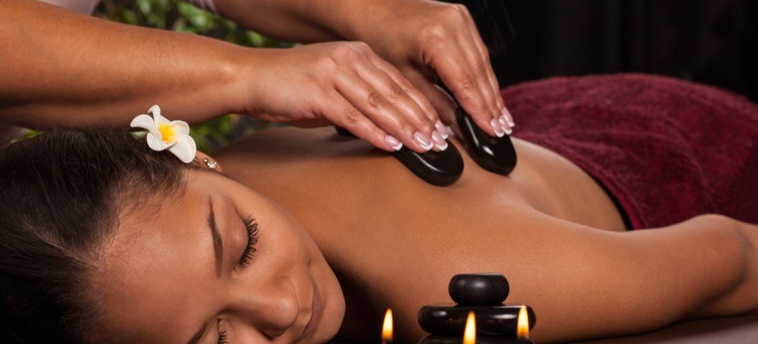 Eine Frau mit geschlossenen Augen liegt entspannt auf einer Massageliege. Sie erhält eine Hot-Stone-Massage auf dem Rücken. Neben ihr brennen kleine farbige Kerzen, im Vordergrund sind dekorative schwarze Steine gestapelt. Die Frau hat eine weiße Blüte im