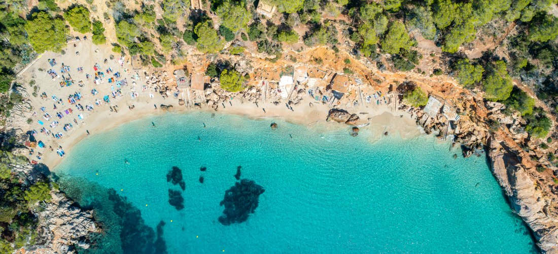 Luftaufnahme der Badebucht Cala Salada auf Ibiza mit türkisblauem Wasser, Sandstrand und umgebenden Pinienwäldern. Am Strand liegen viele Urlauber auf Handtüchern und Liegen.