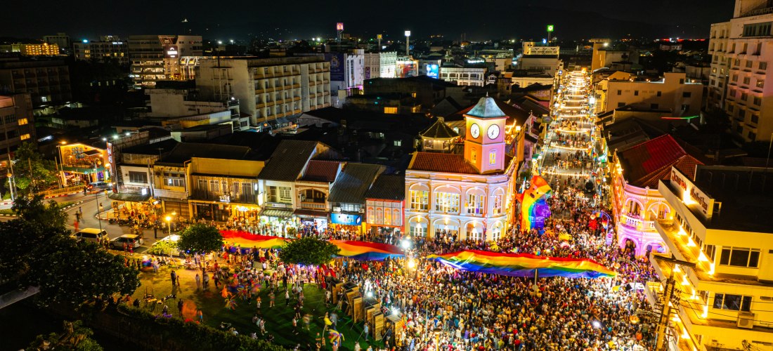 Luftaufnahme des Phuket Pride Festivals bei Nacht. Eine riesige Menschenmenge füllt die geschmückte Straße rund um den ikonischen Uhrenturm. Über dem Festival spannt sich eine große Regenbogenfahne. Die Straße ist hell beleuchtet, Musik und Lichter schaff