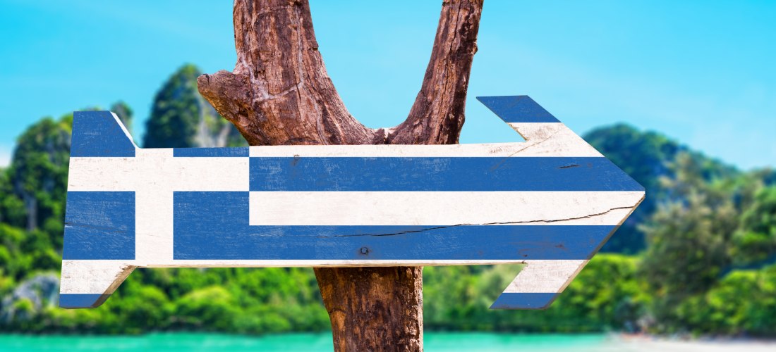 Ein hölzerner Wegweiser in Form eines Pfeils, bemalt mit der griechischen Flagge, ist an einem rustikalen Ast befestigt. Im Hintergrund türkisblaues Wasser, grüne Küste und strahlend blauer Himmel – Symbol für Reise oder Urlaub in Griechenland.