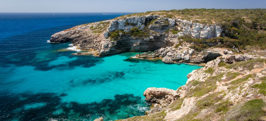 Eine felsige Küstenlandschaft auf Mallorca mit atemberaubend türkisfarbenem Wasser, das sanft in die felsigen Buchten fließt. Die steilen Klippen sind mit mediterraner Vegetation bewachsen und rahmen die Bucht malerisch ein. Das Wasser zeigt unterschiedli