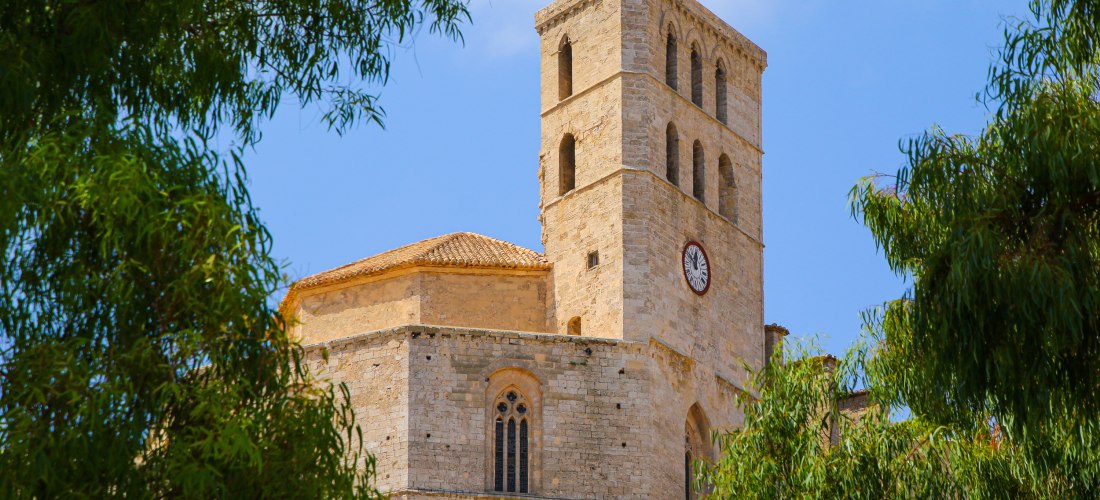 Eine historische Kirche aus Sandstein mit einem rechteckigen Turm, in dem mehrere hohe Rundbogenfenster eingelassen sind. Eine große Uhr ist mittig am Turm angebracht. Das Dach ist mit rötlichen Ziegeln gedeckt, und im Vordergrund rahmen grüne Bäume die S
