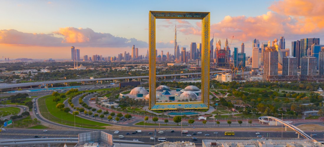 Luftaufnahme des Dubai Frame bei Sonnenuntergang, einem monumentalen, rahmenförmigen Bauwerk aus goldverziertem Metall. Durch den Rahmen ist die Skyline Dubais mit dem Burj Khalifa sichtbar. Darunter liegt ein weitläufiger Park mit Kuppelbauten, Straßen u