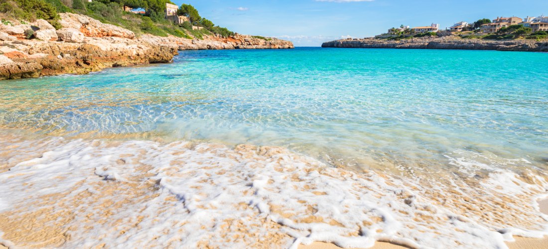 Wellen rollen sanft über den feinen Sandstrand an einer ruhigen Bucht auf Mallorca, mit klarem türkisblauem Wasser und felsiger Küste im Hintergrund