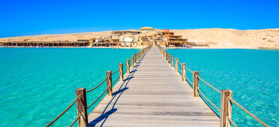 Ein langer hölzerner Steg führt über türkisblaues, kristallklares Wasser auf eine Strandanlage mit strohgedeckten Hütten und Gebäuden in einer wüstenartigen Landschaft zu. Der Himmel ist strahlend blau und wolkenlos.