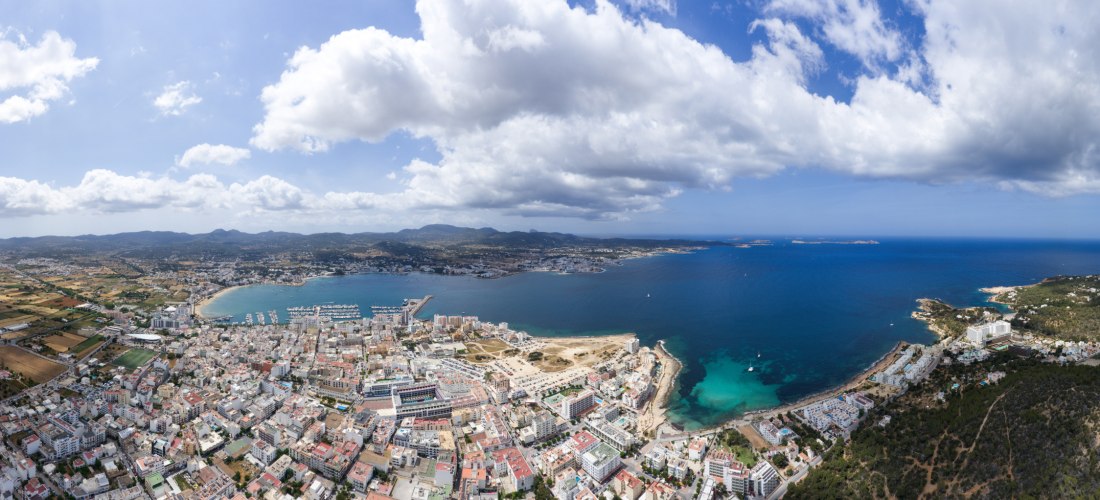 Luftaufnahme von Sant Antoni de Portmany auf Ibiza: Stadt am Meer mit großer Bucht, Marina und Mole; rechts türkisfarbene Buchten, im Hinterland Felder und grüne Hügel unter teils bewölktem Himmel.