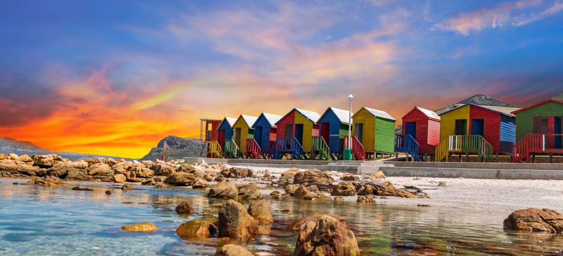 Reihe bunter viktorianischer Strandhütten am Muizenberg Beach in Kapstadt, Südafrika. Die Hütten stehen auf Stelzen direkt am Strand und leuchten in kräftigen Farben wie Rot, Blau, Gelb und Grün. Im Vordergrund glasklares, flaches Wasser mit Felsen und Ki