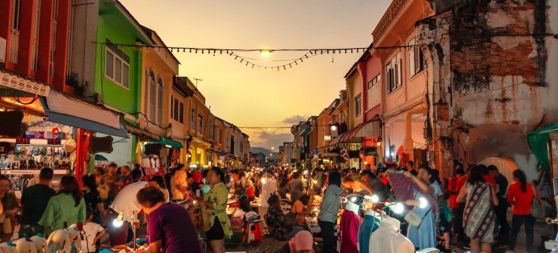 Menschenmengen schlendern über einen lebhaften Nachtmarkt in Phuket Old Town bei Sonnenuntergang. Bunte Kolonialgebäude säumen die Straße, Verkaufsstände bieten Kleidung, Souvenirs und Streetfood an. Warme Lichter hängen über der Straße und sorgen für sti