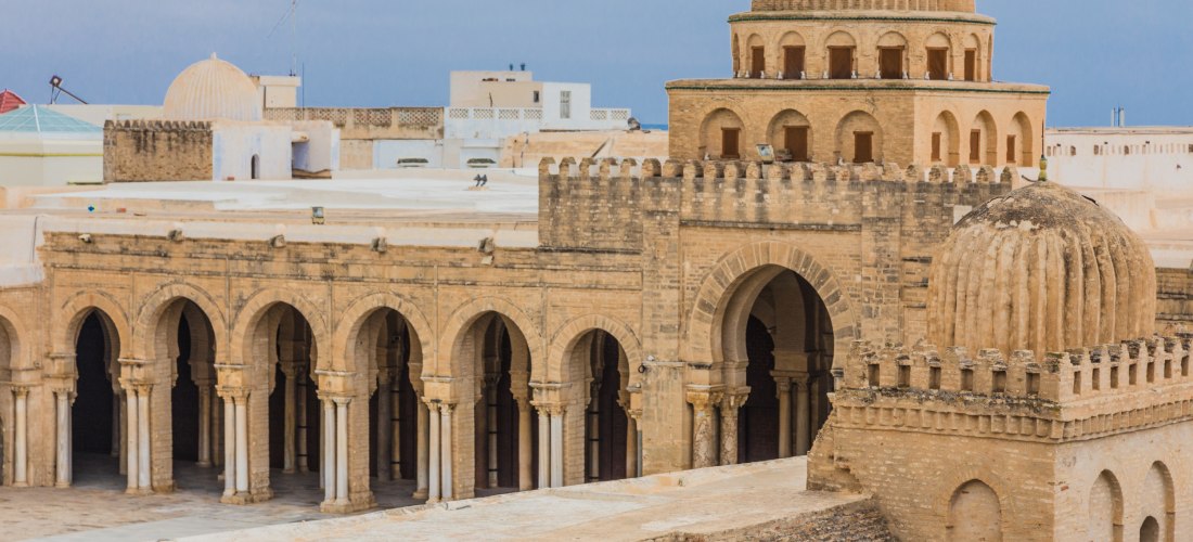 Die imposante Große Moschee von Kairouan in Tunesien. Das Foto zeigt die monumentale Architektur mit ihren massiven Torbögen, kunstvollen Kuppeln und detailreichen Mauern. Die Moschee thront majestätisch inmitten traditioneller Gebäude und strahlt eine be