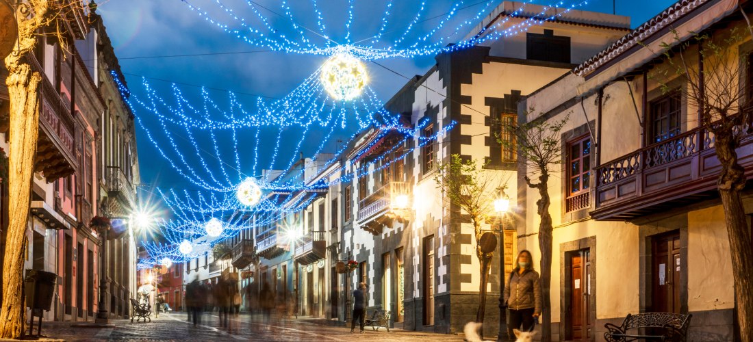 Straße in Teror auf Gran Canaria am Abend, festlich geschmückt mit leuchtenden Lichterketten, Menschen spazieren entlang der traditionellen Häuserfassaden, im Vordergrund ein Hund.