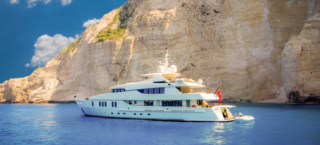 >   Das Bild zeigt eine große, moderne Luxus-Yacht, die ruhig auf dem tiefblauen Wasser liegt.    Im Hintergrund erhebt sich eine gewaltige Steilküste aus hellem Sandstein, durchzogen von Rillen und leichten Farbunterschieden.   Auf dem Deck der Yacht bef