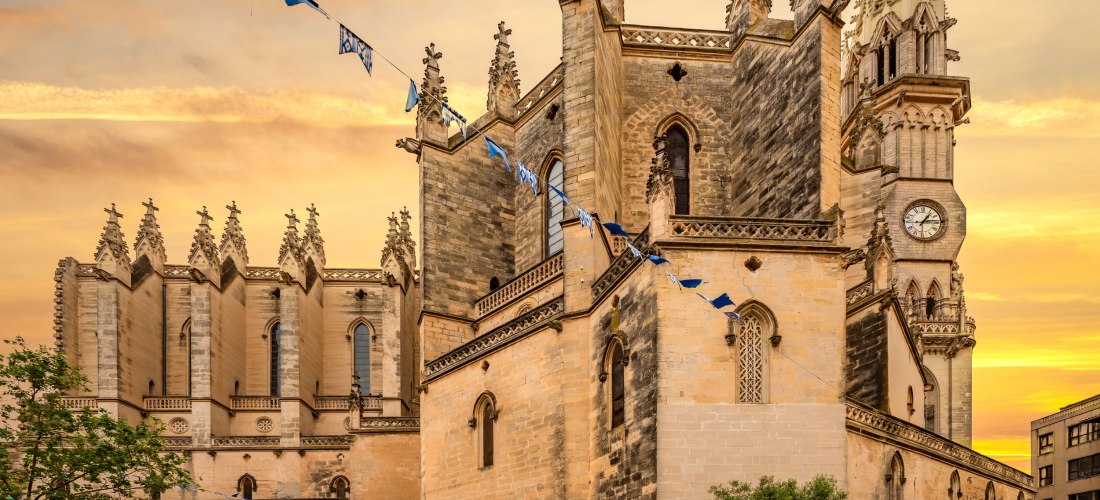 Eine beeindruckende Kirche im neugotischen Stil, wahrscheinlich in Spanien oder Südfrankreich, mit einer Fassade aus hellem Sandstein. Die Kirche hat viele spitze Türme und Zinnen, die an eine Burg erinnern. Blaue und weiße Fahnen sind an einem Seil zwisc