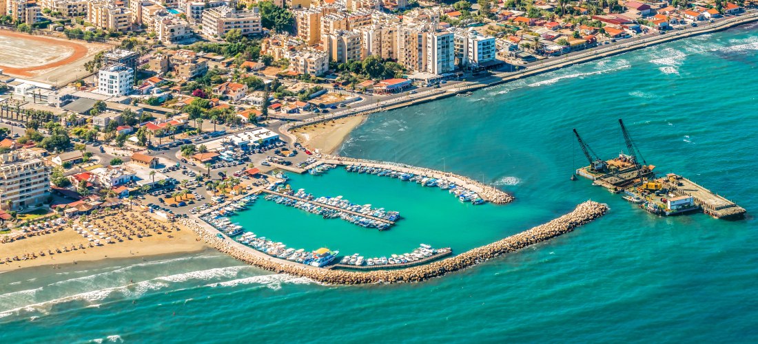 Larnaca Marina aus der Luft: halbkreisförmiges Hafenbecken mit Yachten, Strandabschnitt und Stadtpanorama an Zyperns Südküste.
