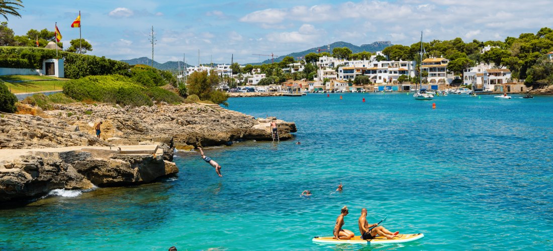 Badegäste im türkisblauen Wasser von Cala d’Or auf Mallorca, mit Felsen, Klippenspringern und Stand-Up-Paddlern vor weißen Häusern am Ufer