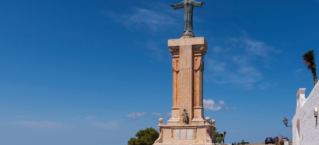Christusstatue mit ausgebreiteten Armen auf einem hohen Steinsockel auf dem Monte Toro, dem höchsten Punkt Menorcas. Die Statue steht auf einem Aussichtsplateau mit Blick über die Insel. Der Himmel ist klar blau, es sind nur wenige kleine Wolken zu sehen.