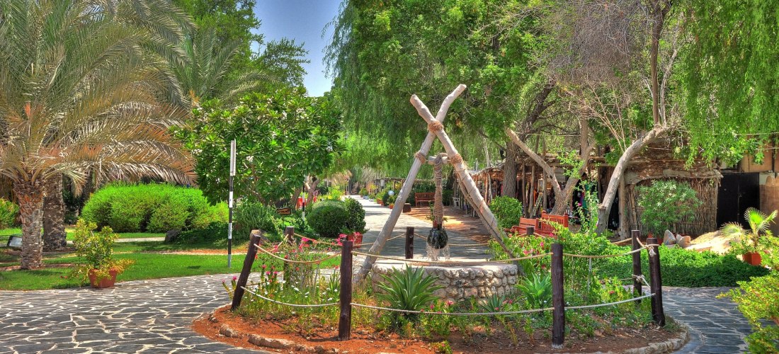 Traditionelles Heritage Village in Abu Dhabi mit Palmen, grünen Bäumen, Steingartenwegen und dekorativem Brunnen inmitten einer parkähnlichen Anlage