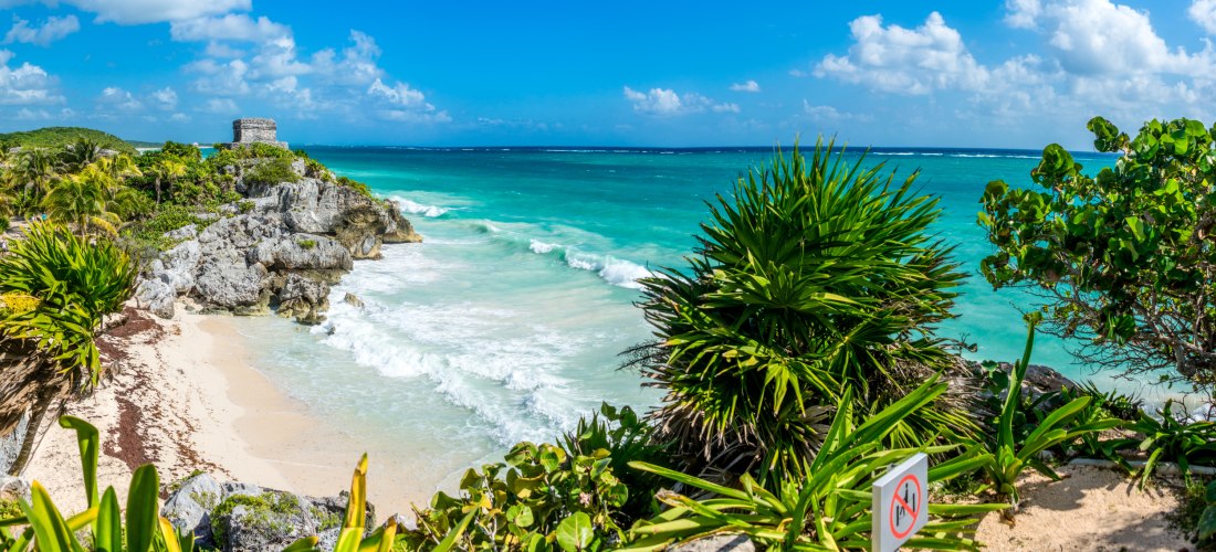Panoramaansicht der Ruinen von Tulum auf einer Klippe mit tropischer Vegetation, türkisblauem Meer und Wellen, die gegen den weißen Sandstrand schlagen