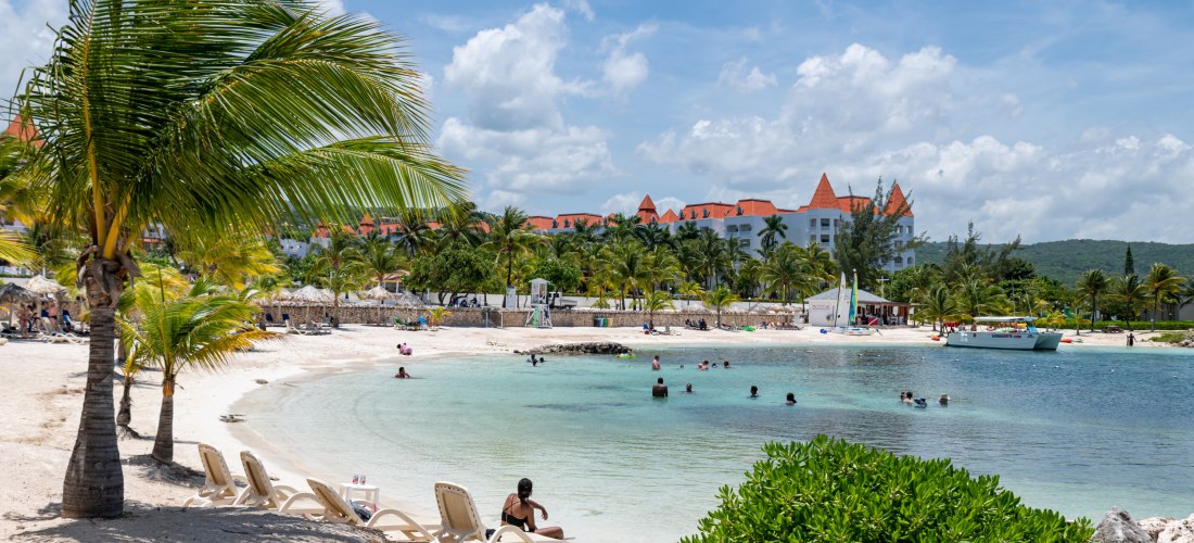 Karibischer Strand mit Sonnenliegen, Palmen und türkisfarbenem Wasser – Urlauber genießen das tropische Klima vor einer Resortanlage.
