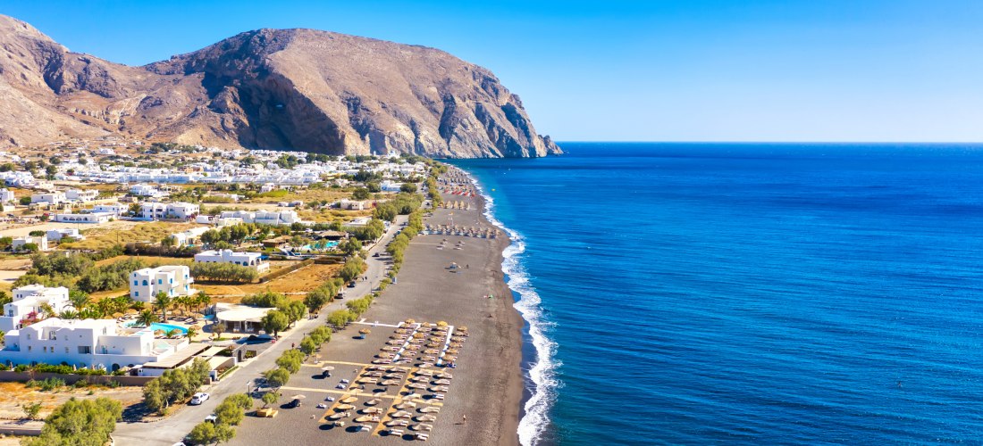 Luftaufnahme vom Kamari Beach auf Santorini, mit einem langen dunklen Sandstrand, der sich entlang der Küste erstreckt. Weiße Häuser mit Pools im traditionellen kykladischen Stil verteilen sich im Hintergrund vor einer steilen Felskulisse. Das Meer leucht