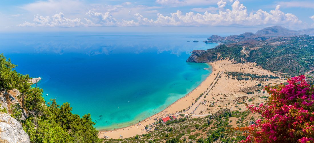 Das Bild zeigt eine weite Panoramansicht auf eine atemberaubende Küstenlandschaft auf einer griechischen Insel – wahrscheinlich Kreta oder Rhodos.    Im Vordergrund wächst eine leuchtend pinke Bougainvillea. Rechts unten beginnt ein goldgelber Sandstrand,