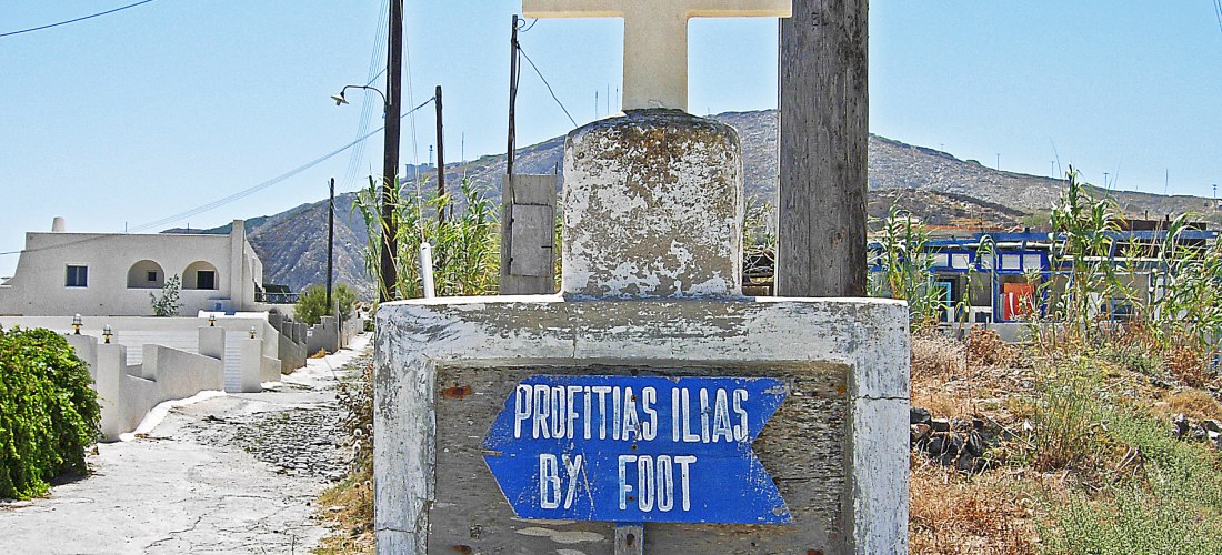 Ein verwitterter Wegweiser aus Beton mit einem weißen Kreuz obenauf. Darunter zeigt ein blauer Pfeil mit weißer Schrift 'Profitis Ilias by foot' nach rechts. Der Weg führt durch eine trockene, leicht bewachsene Landschaft mit Stromleitungen und weißen Häu