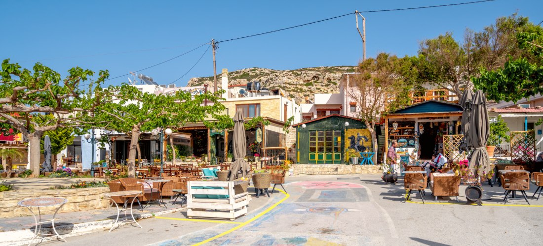 Ein kleiner, bunter Platz im Ort Matala auf Kreta mit Tavernen, Cafés und kleinen Läden. Im Vordergrund stehen Tische und Stühle unter schattigen Bäumen, im Hintergrund bunte Hausfassaden und ein Souvenirladen. Der Himmel ist klar und blau, das Licht warm
