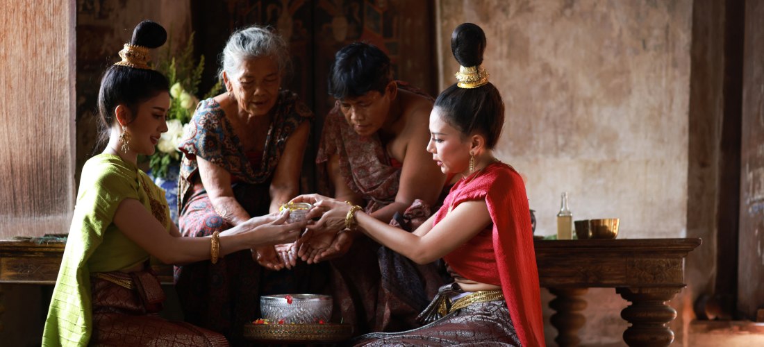 Zwei junge Frauen in traditioneller thailändischer Kleidung gießen Wasser über die Hände zweier älterer Menschen. Alle sitzen respektvoll am Boden in einem historischen Raum. Eine silberne Schale mit Blüten steht vor ihnen.