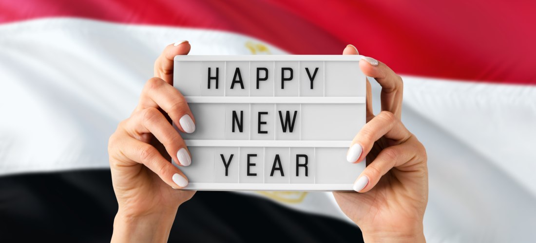 Zwei Hände halten ein Leuchtschild mit der Aufschrift 'HAPPY NEW YEAR' vor einer wehenden ägyptischen Flagge in Rot, Weiß und Schwarz.