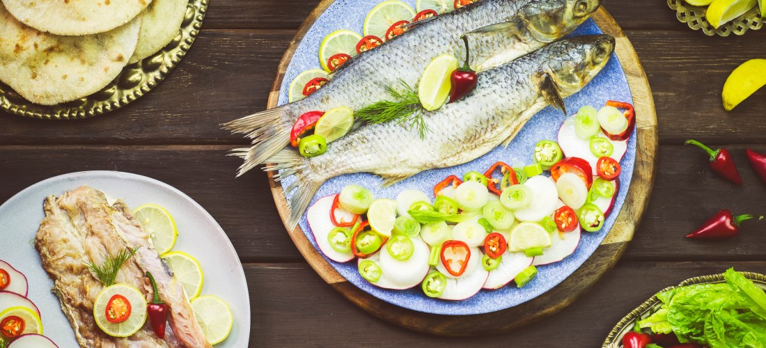 Zwei rohe Fische auf einem blauen Teller, garniert mit Limettenscheiben, Chili, Frühlingszwiebeln und Dill. Umgeben von Fladenbrot, Hummus, Gemüse und Zitronen auf einem Holztisch.