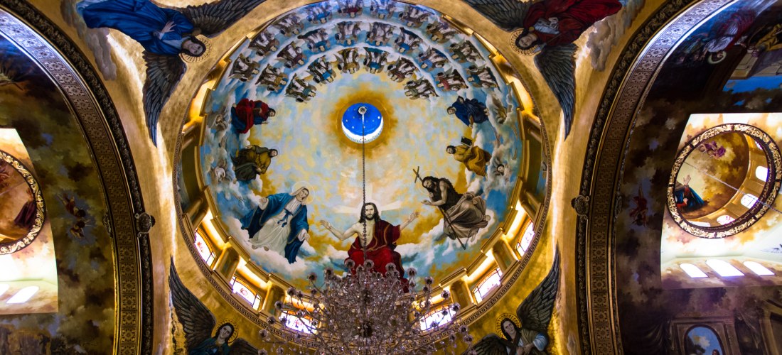 Blick in eine bemalte Kirchenkuppel mit einer Darstellung Christi in der Mitte, umgeben von Maria, Johannes dem Täufer und zahlreichen Heiligenfiguren sowie Engeln. Die Szene ist himmlisch inszeniert, mit goldener Umrahmung und detailreichen Fresken. Ein 