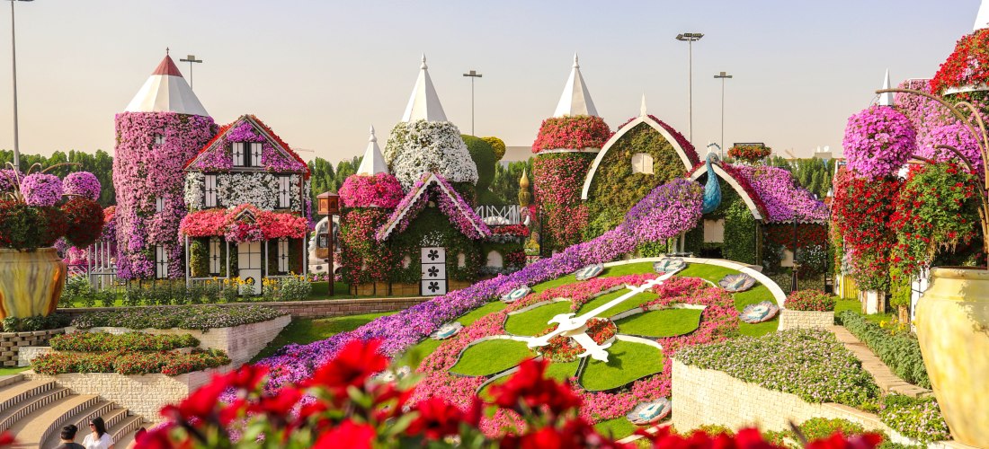 Märchenhafte Blumeninstallation im Dubai Miracle Garden mit kunstvoll dekorierten Häuschen und Türmen, die komplett mit blühenden Pflanzen in Pink, Lila, Weiß und Rot bedeckt sind. Im Vordergrund blühen rote Petunien, rechts ein übergroßer Blumentopf. Auf
