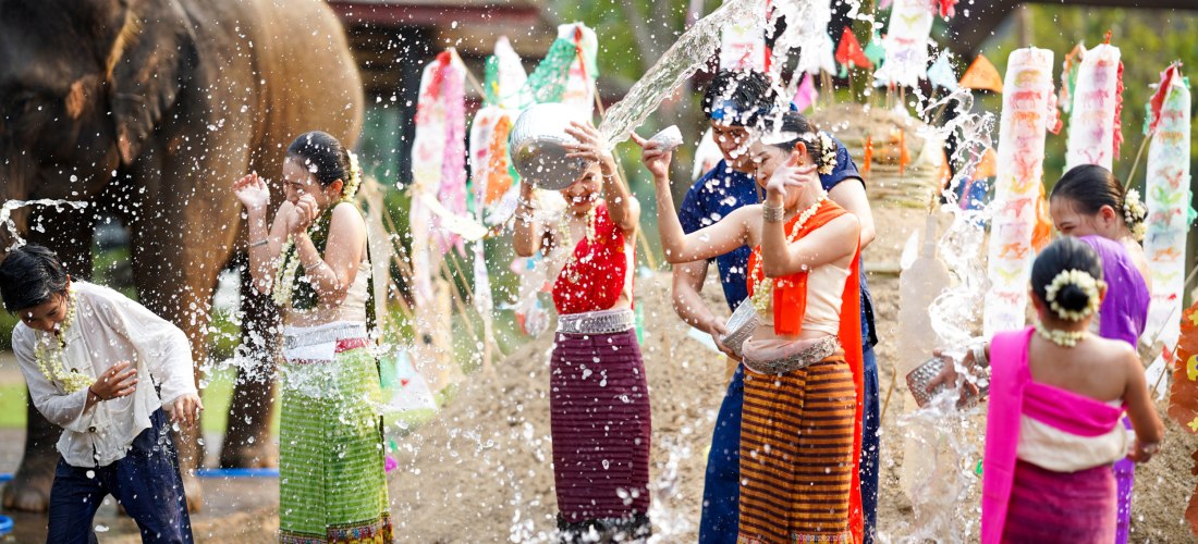Das Bild zeigt eine lebhafte Szene des thailändischen Wasserfestes Songkran. Männer, Frauen und Kinder in farbenfroher, traditioneller Kleidung lachen und spritzen sich gegenseitig mit Wasser aus Schalen und Gefäßen. Wasser fliegt durch die Luft und sorgt