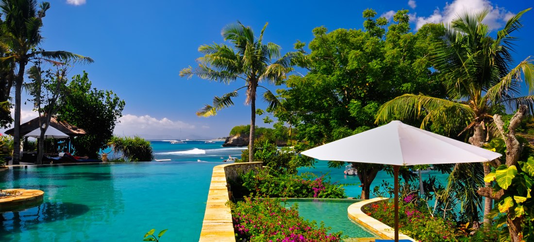 Blick auf einen Infinity-Pool in einer tropischen Anlage auf Bali. Umgeben von Palmen, Blumen und einem weißen Sonnenschirm erstreckt sich der türkisfarbene Pool bis zum Meer. Im Hintergrund sieht man eine kleine Bucht mit Booten und eine sattgrüne Küste 