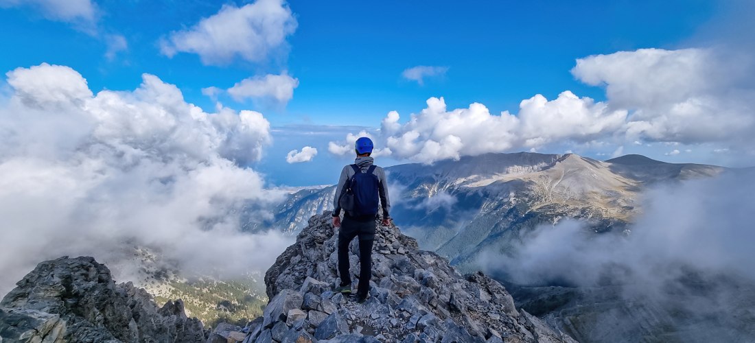 Bergsteiger mit Helm und Rucksack auf einem Gipfel des Olymps, umgeben von Wolken und Blick auf die umliegenden Berge.