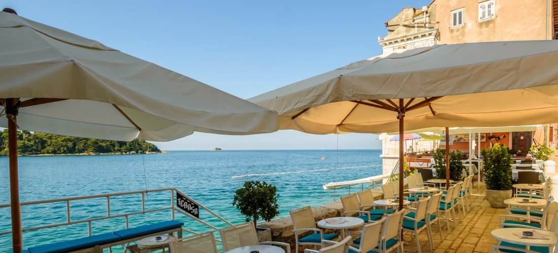 Kroatien - Rovinj Gemütliche Restaurantterrasse mit weißen Stühlen und Sonnenschirmen direkt am Wasser in Rovinj, Kroatien. Blick auf das türkisfarbene Meer und eine kleine Insel am Horizont.