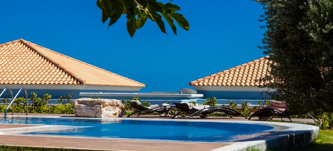 Das Bild zeigt einen gepflegten Poolbereich in einer modernen Hotelanlage.    Der runde Pool ist von einer Holzterrasse umgeben, auf der mehrere elegante Liegestühle mit weißen Handtüchern bereitstehen.    Im Hintergrund sind zwei Gebäude mit typischen Te