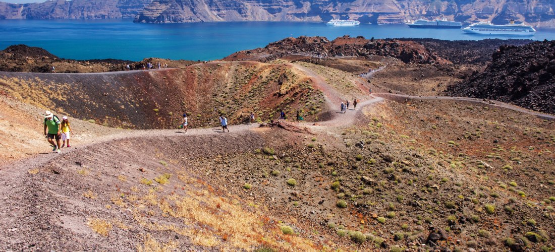 Mehrere Wanderer gehen auf einem kurvigen Pfad über die karge, rotbraune Vulkanlandschaft der Insel Nea Kameni. Im Hintergrund ist das tiefblaue Meer zu sehen, dahinter erhebt sich steil die Insel Santorini mit ihren weißen Häusern, die auf der Felskante 