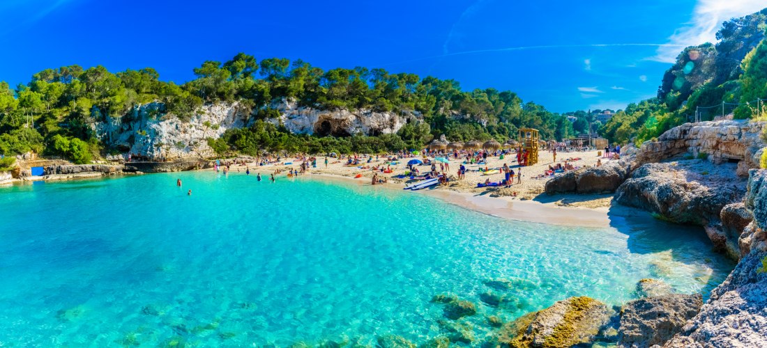 Menschen entspannen sich am malerischen Strand Cala auf Mallorca mit kristallklarem Wasser und Pinienwald im Hintergrund