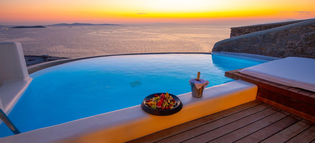Infinity-Pool auf einer Terrasse mit Blick auf das ruhige Meer bei Sonnenuntergang. Auf dem Rand des Pools stehen ein Teller mit frischem Obst und ein Eiskübel mit einer Flasche Sekt. Der Himmel leuchtet in warmen Gelb- und Orangetönen.